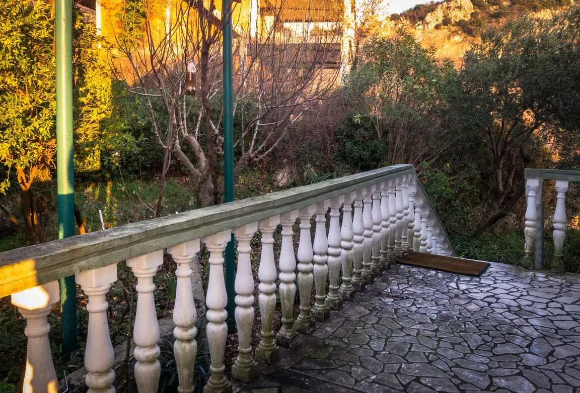 Terasa sa kamenim podom i ogradom od betonskih stubića okružena zelenilom.
