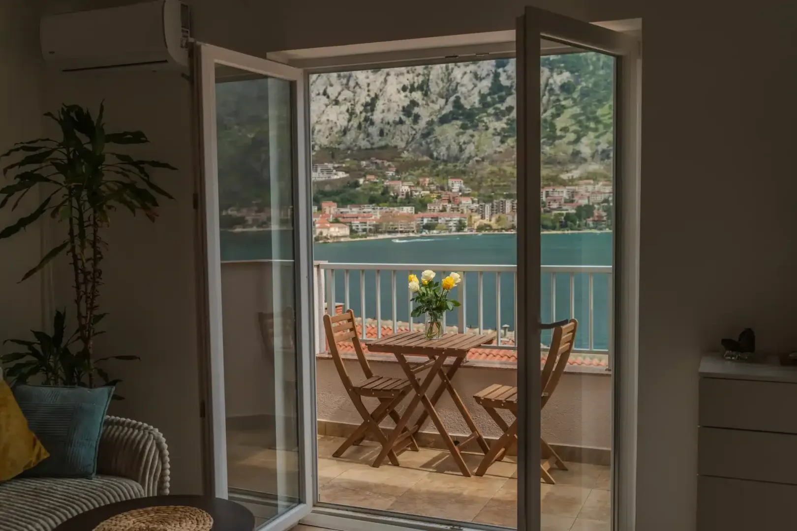 Pogled kroz otvorena balkonska vrata na terasu sa garniturom i more.