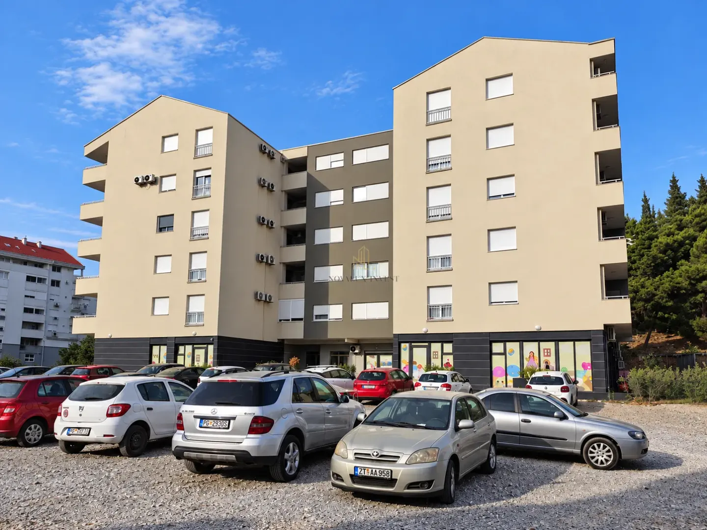 Pogled na spoljašnjost stambene zgrade sa parkingom u prvom planu.