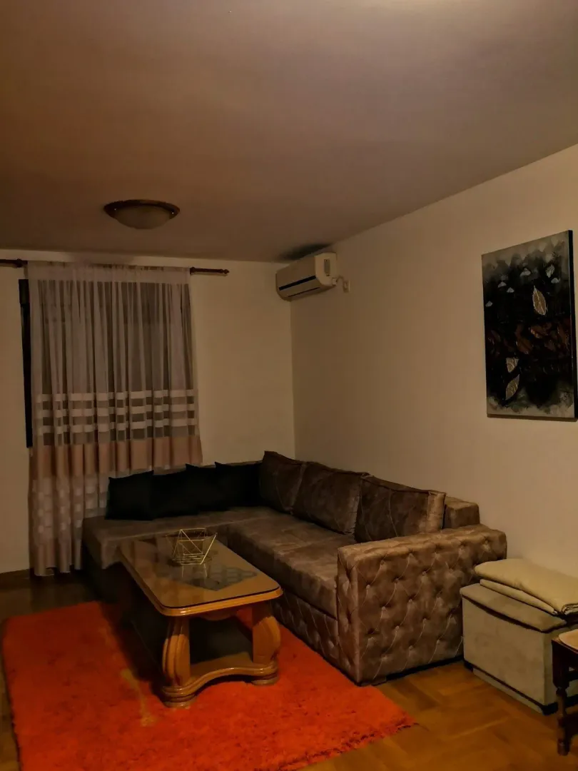 Dnevna soba sa ugaonom garniturom, drvenim klub stolom i narandžastim tepihom.