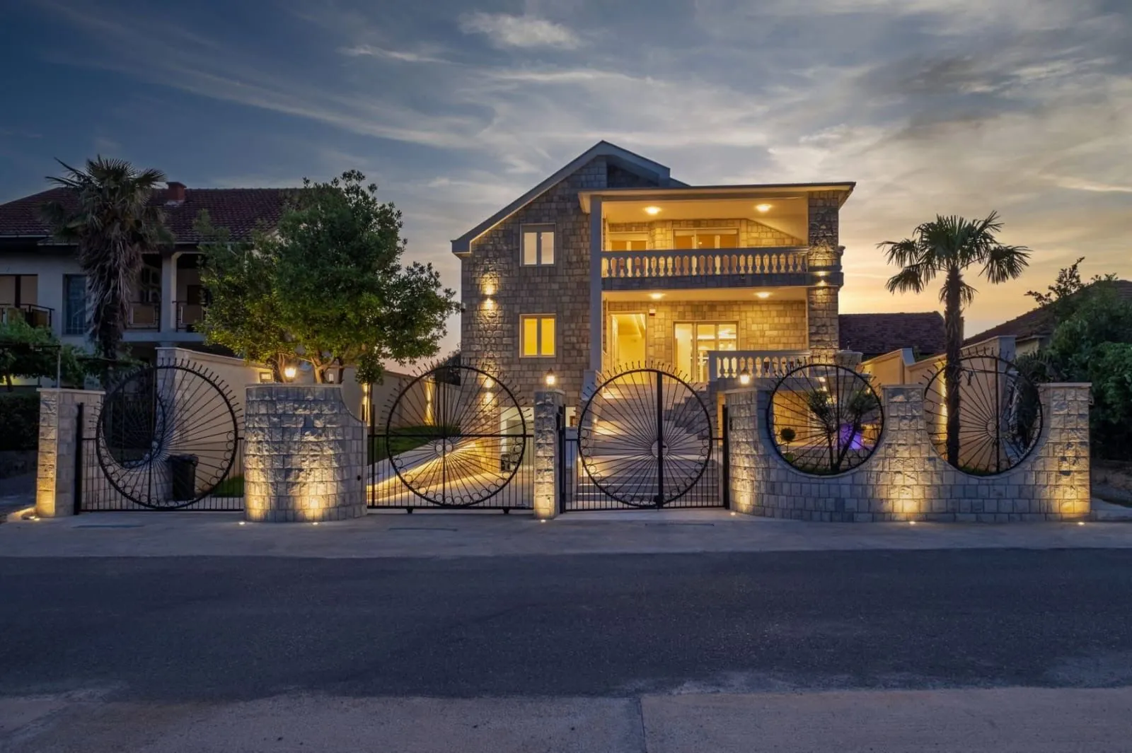 View of the illuminated stone house facade with a landscaped yard and fence at dusk.