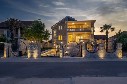 View of the illuminated stone house facade with a landscaped yard and fence at dusk. - image 14
