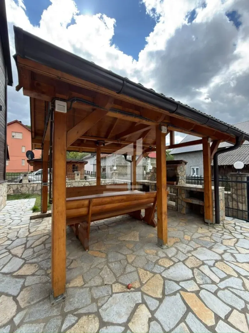 Drvena sjenica sa garniturom za sjedenje i kamenim roštiljem u dvorištu.