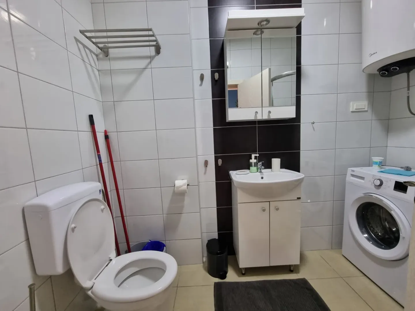 Kupatilo opremljeno sanitarijama, umivaonikom, veš mašinom i bijelim pločicama.