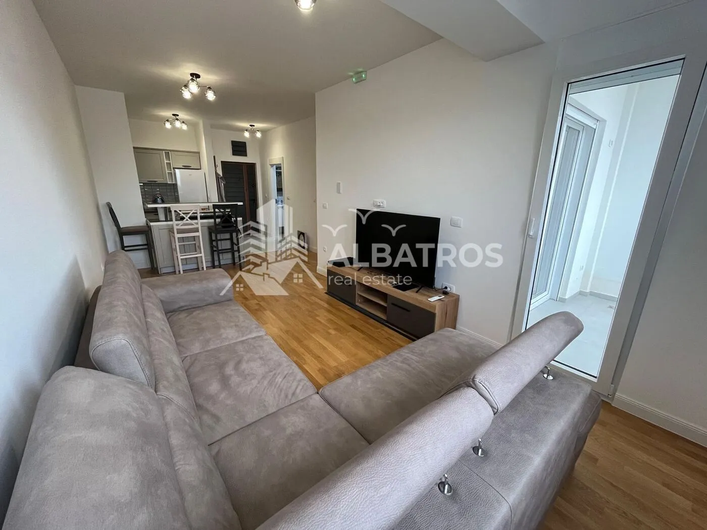 Living room with a sectional sofa, TV unit, and a view of the kitchen in the background.