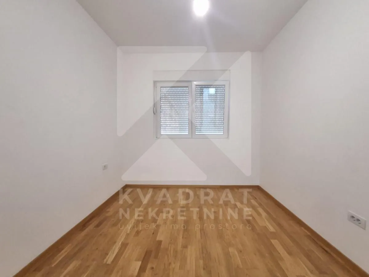 Prazna soba sa bijelim zidovima, drvenim parketom i prozorom.