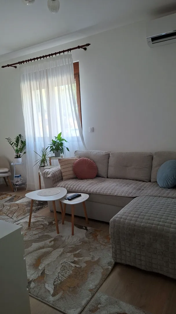 Dnevna soba sa garniturom, stočićem i prozorom sa bijelim zavjesama.