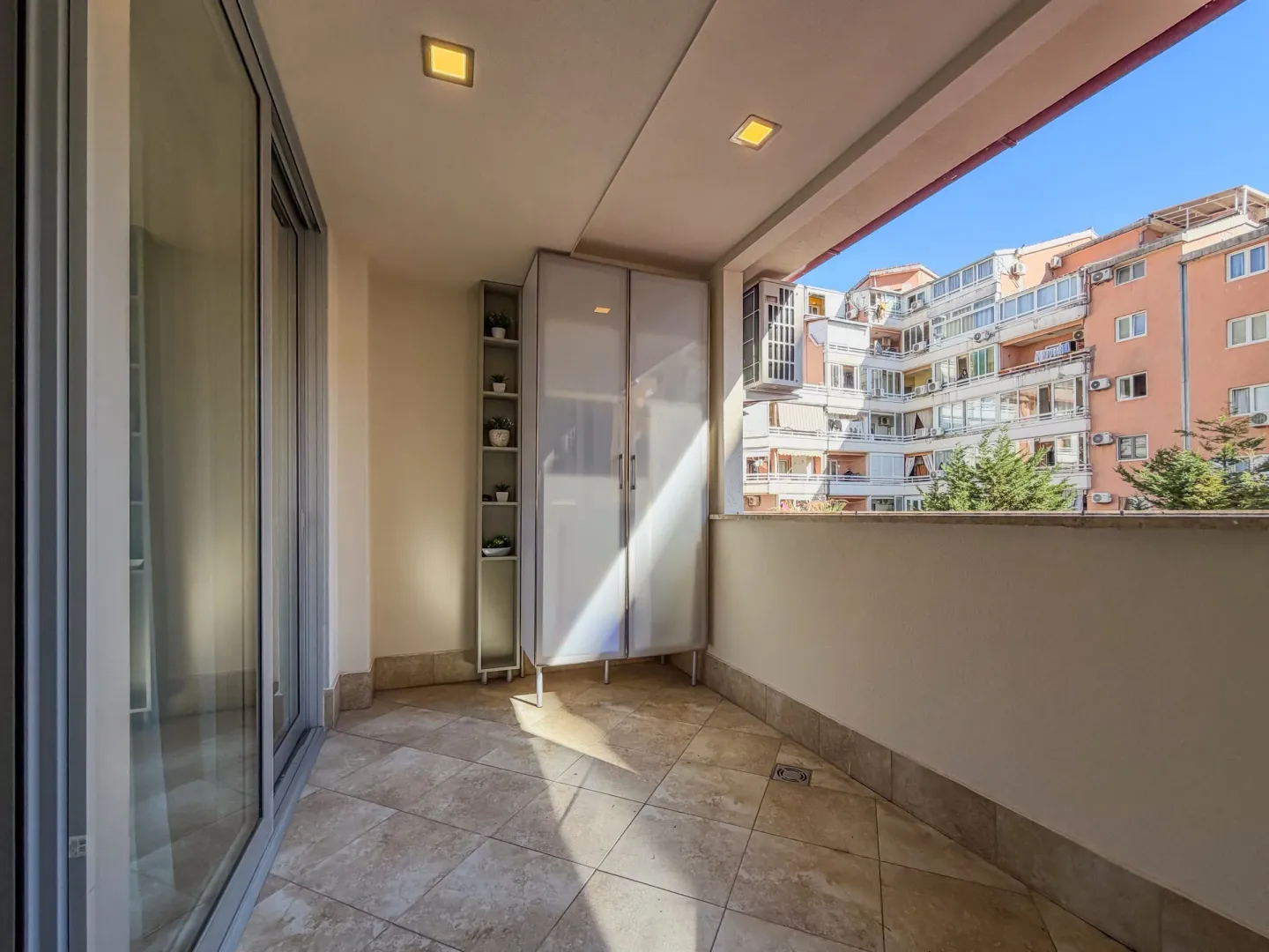 Natkriveni balkon sa pločicama i ugradnim ormarom, sa pogledom na okolne zgrade.