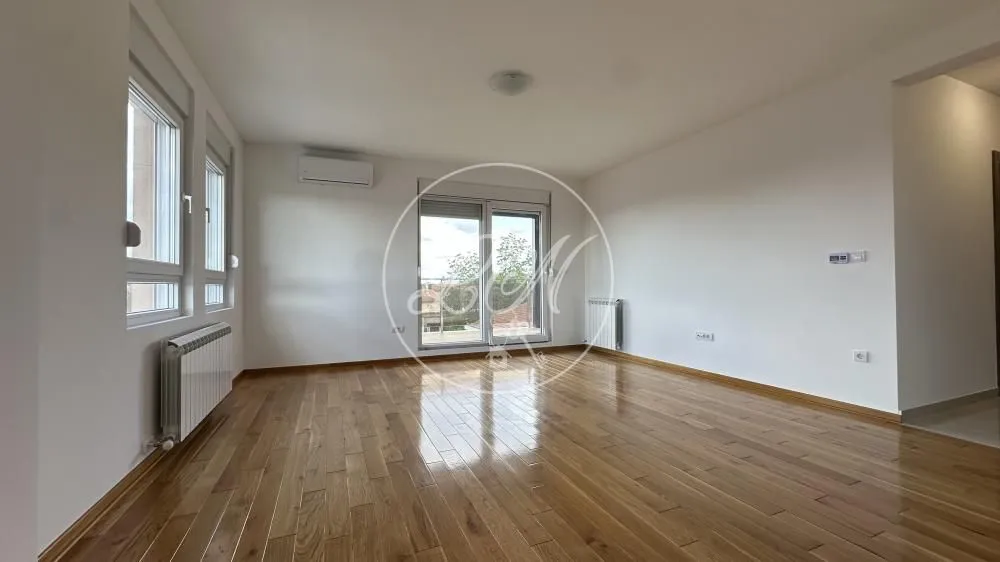 Prazna soba sa drvenim podom, belim zidovima i prozorima.