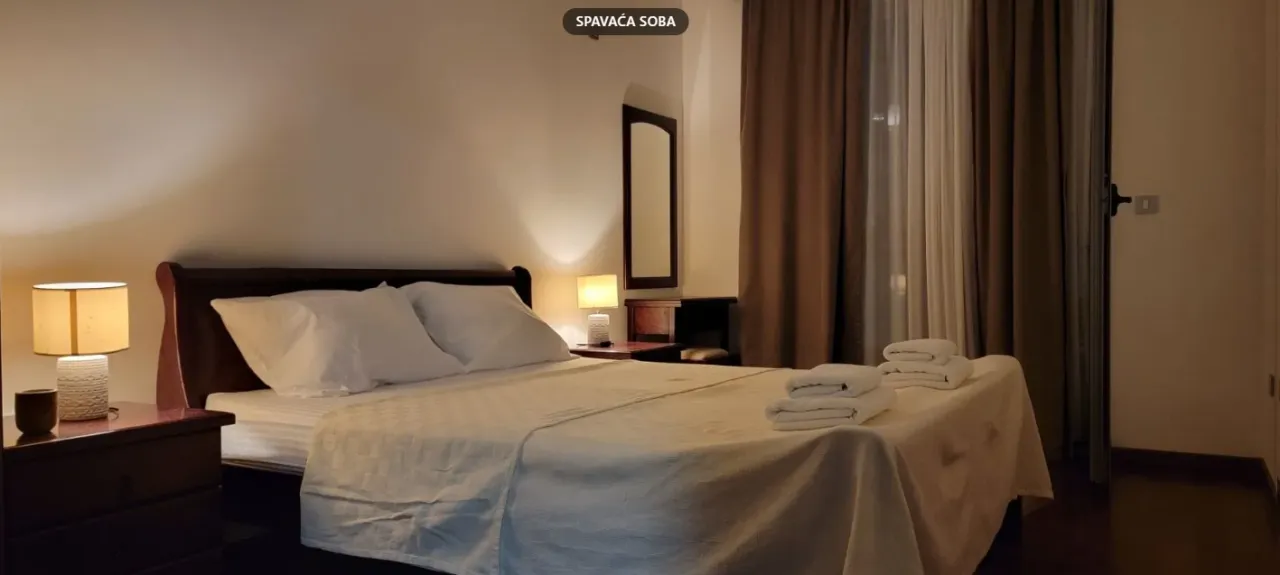 Namještena spavaća soba sa bračnim krevetom, noćnim stočićima i zavjesama.
