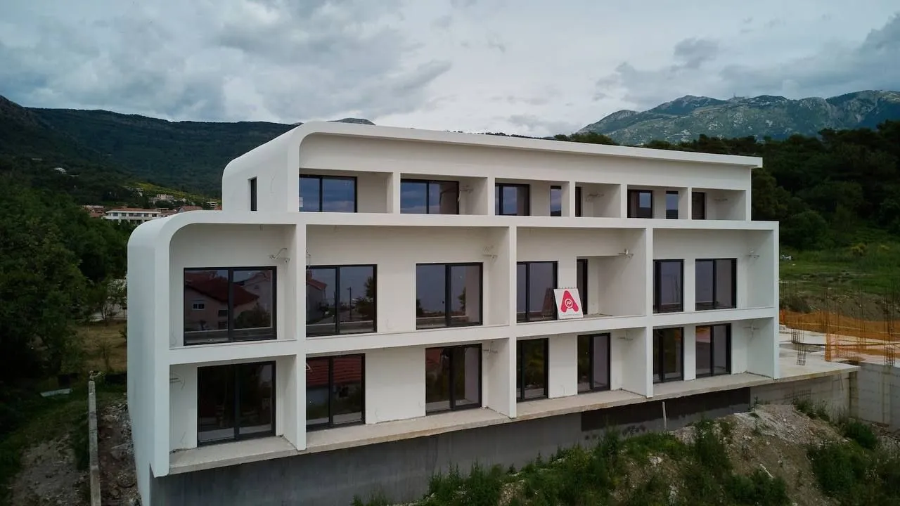 Pogled iz vazduha na modernu bijelu fasadu zgrade sa terasama i planinskim pejzažom u pozadini.