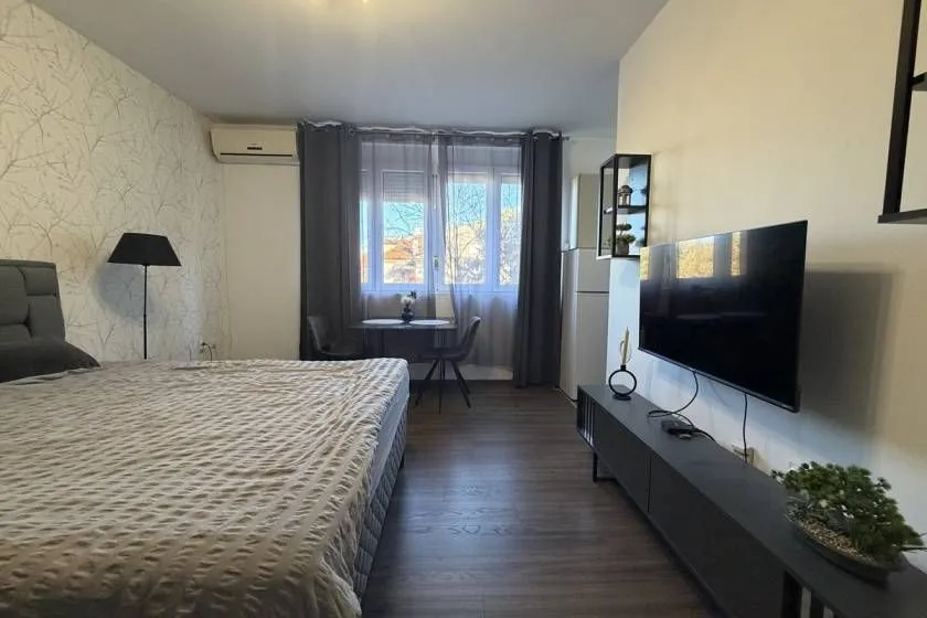 Nameštena spavaća soba sa bračnim krevetom, televizorom i trpezarijskim stolom pored prozora.