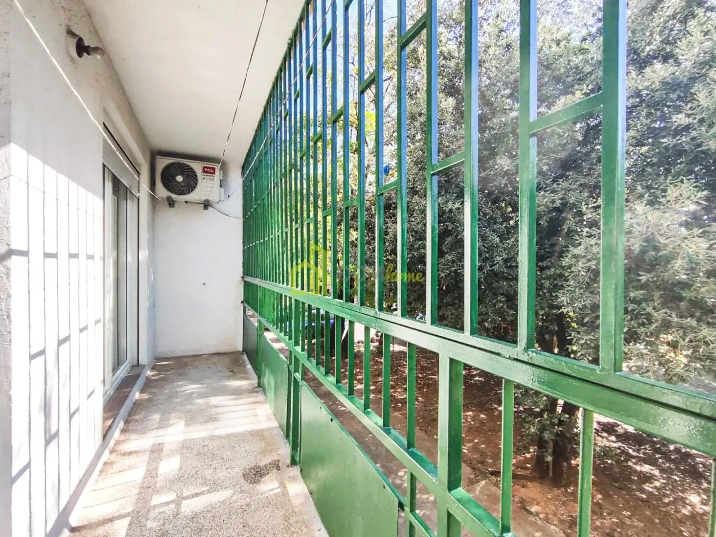 Dugačak balkon sa zelenom zaštitnom ogradom i pogledom na drveće.