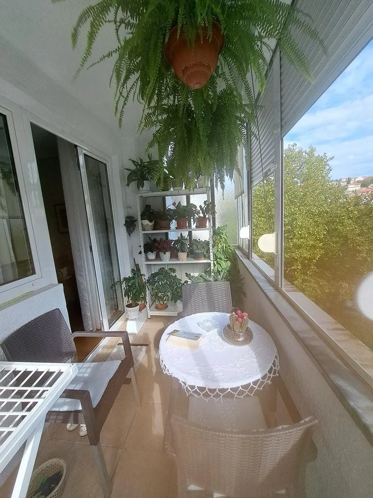 Zastakljeni balkon sa garniturom za sjedenje i mnoštvom biljaka.