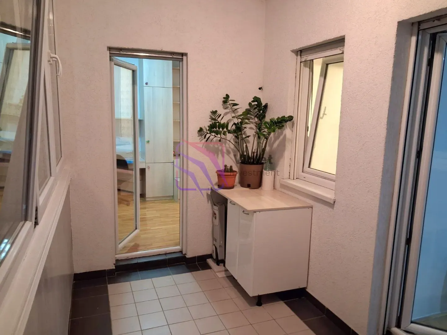 Zastakljeni balkon sa pločicama, komodom i saksijama sa cvijećem.