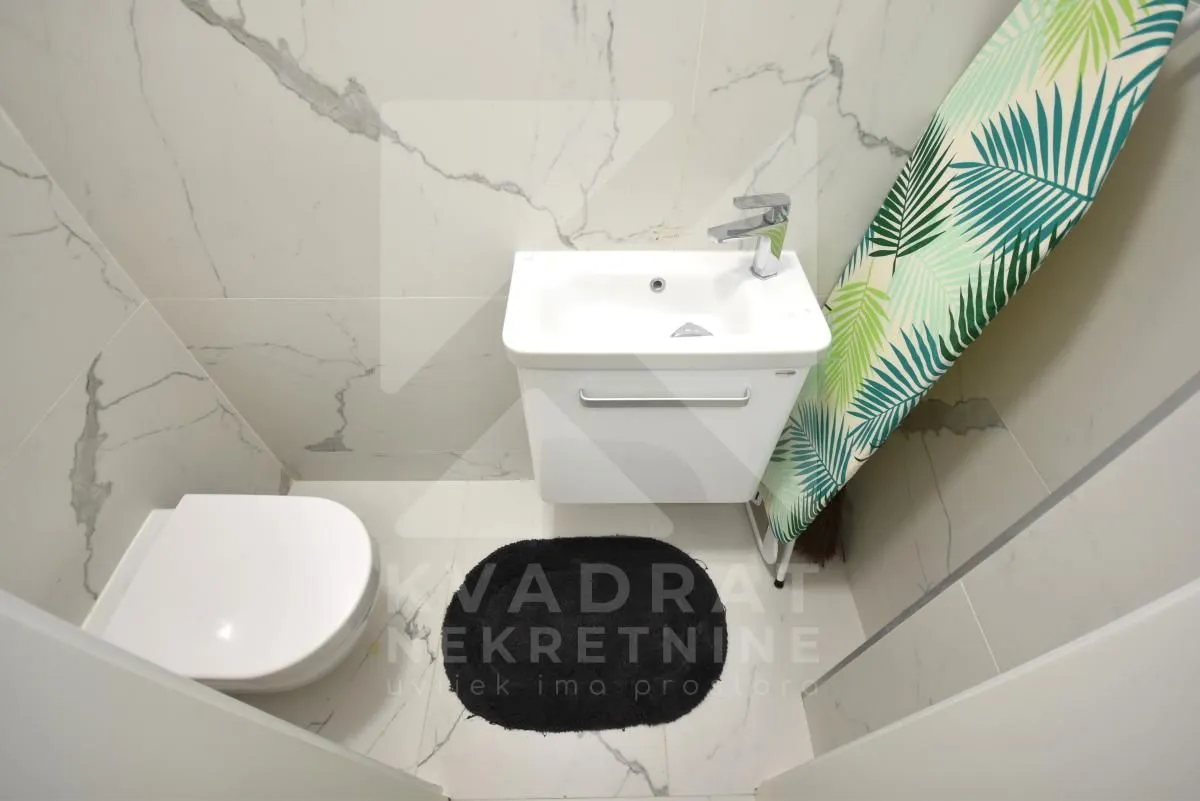Smaller bathroom lined with marble tiles containing a toilet, washbasin, and ironing board.