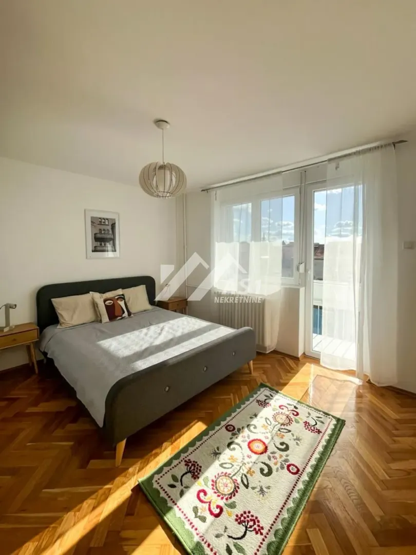 Nameštena spavaća soba sa bračnim krevetom, parketom i izlazom na balkon.