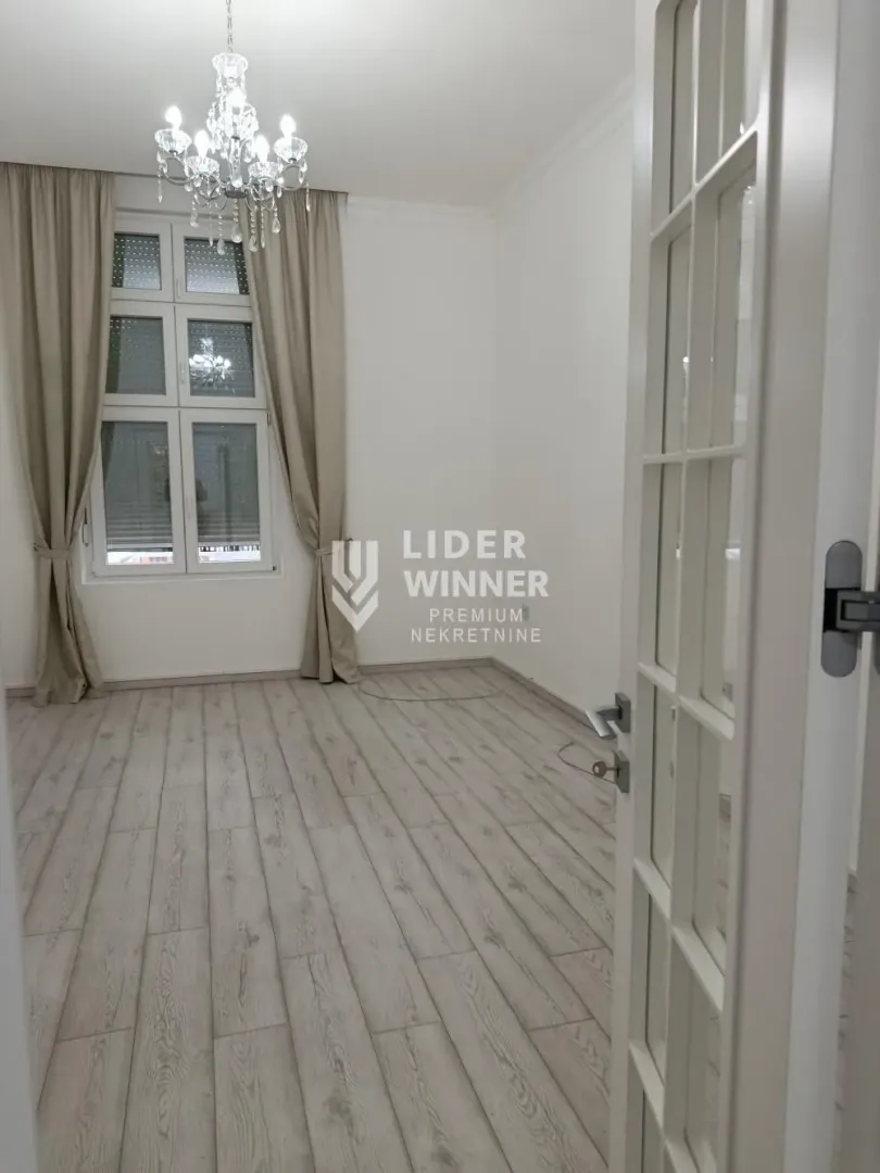 Prazna soba sa svetlim laminatom, prozorom i lusterom, gledana kroz otvorena vrata.