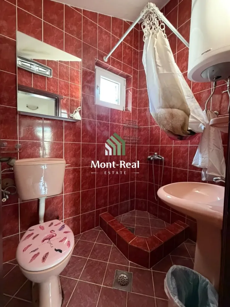 Kupatilo obloženo crvenim pločicama sa wc šoljom, lavaboom i tuš kabinom.