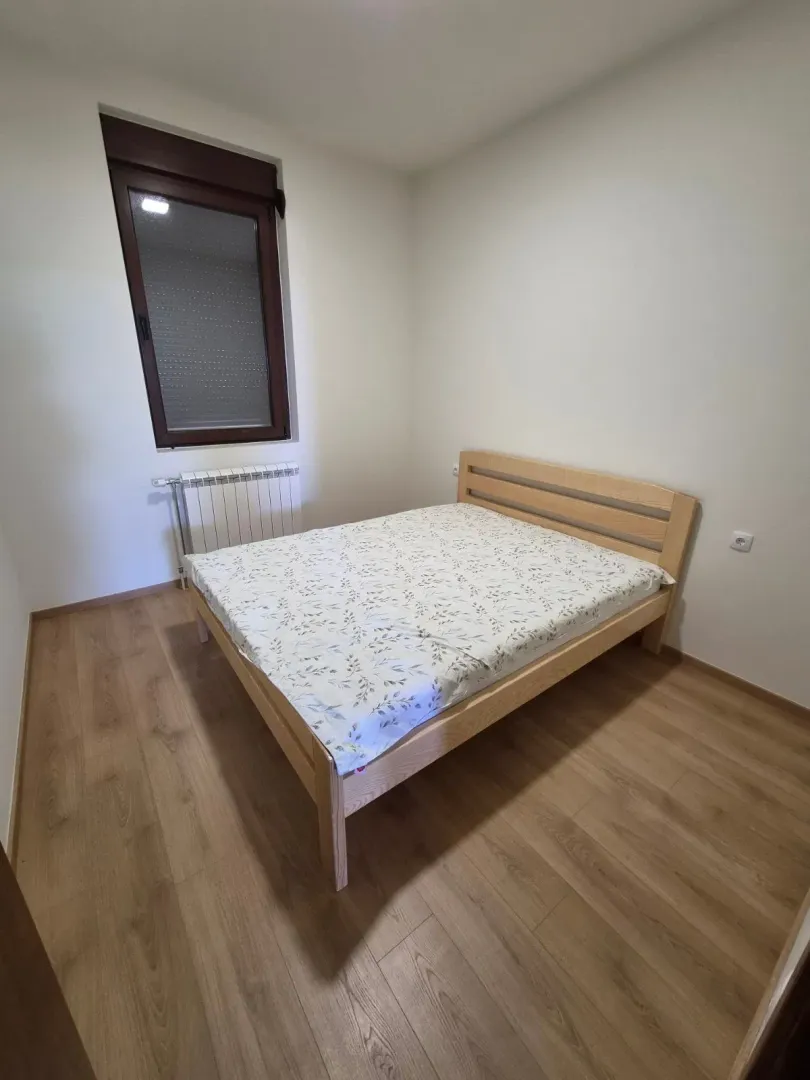 Manja spavaća soba sa bračnim krevetom, drvenim podom i prozorom.