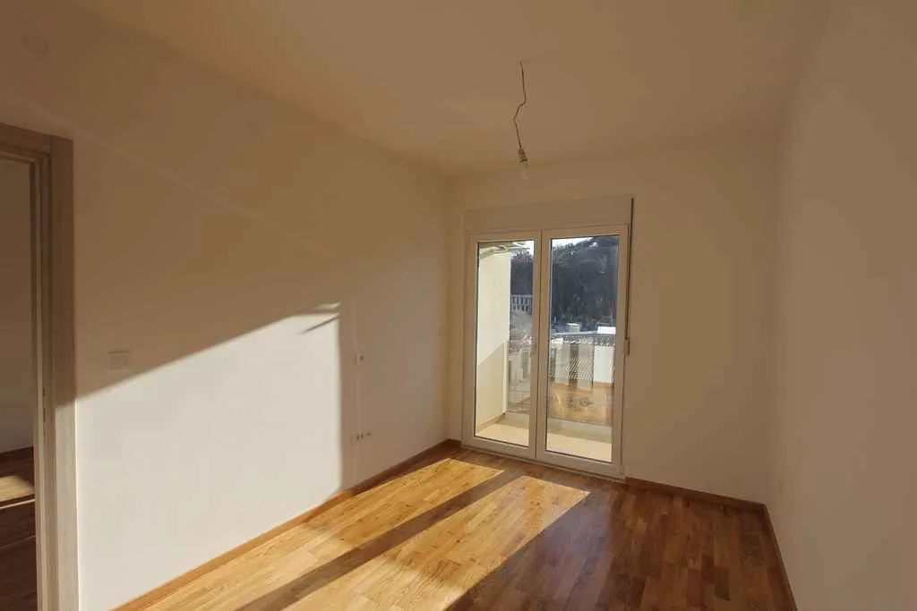 Prazna, svijetla soba sa drvenim podom i izlazom na balkon.