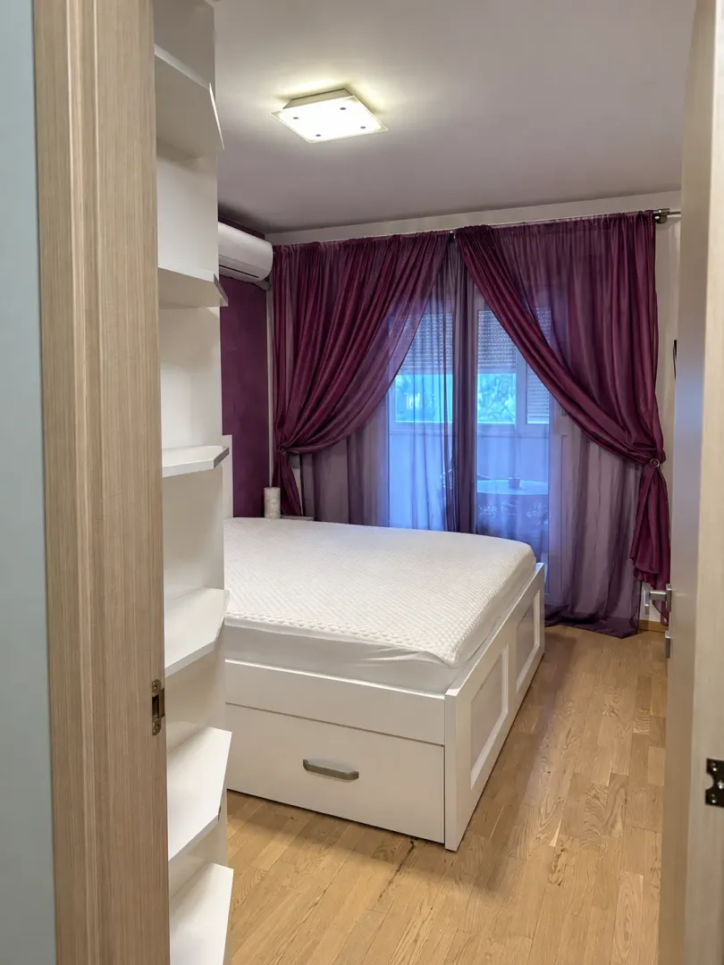 Pogled iz hodnika na spavaću sobu sa bijelim krevetom i ljubičastim zavjesama.