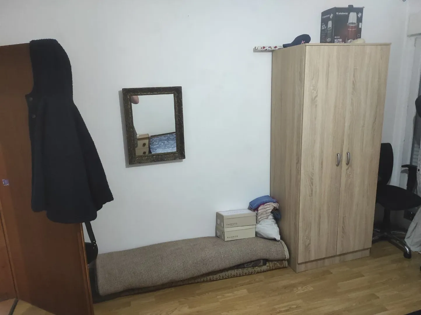 A simple room featuring a wooden wardrobe, a wall-mounted mirror, and parquet flooring.