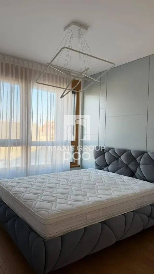 Spavaća soba sa bračnim krevetom, modernim lusterom i velikim prozorom sa zavesama.