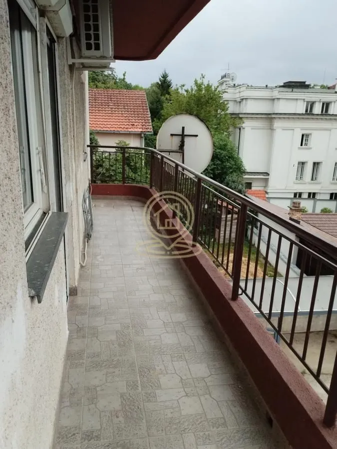Dugi popločani balkon sa metalnom ogradom i pogledom na okolne zgrade.