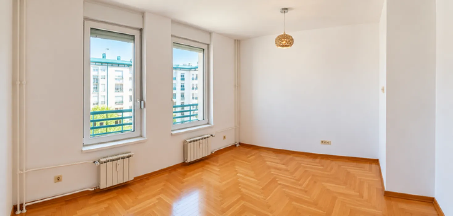 Empty bright room with hardwood flooring and two windows overlooking neighboring buildings.