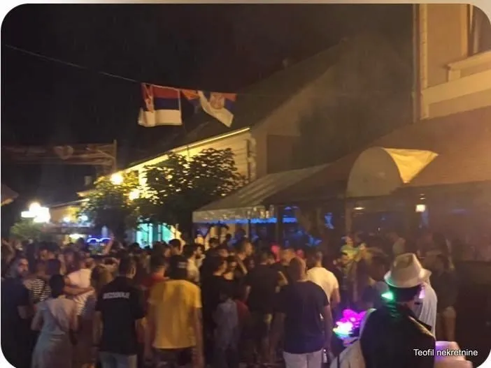 Noćni prikaz ulice sa okupljenim ljudima ispred ugostiteljskog objekta.