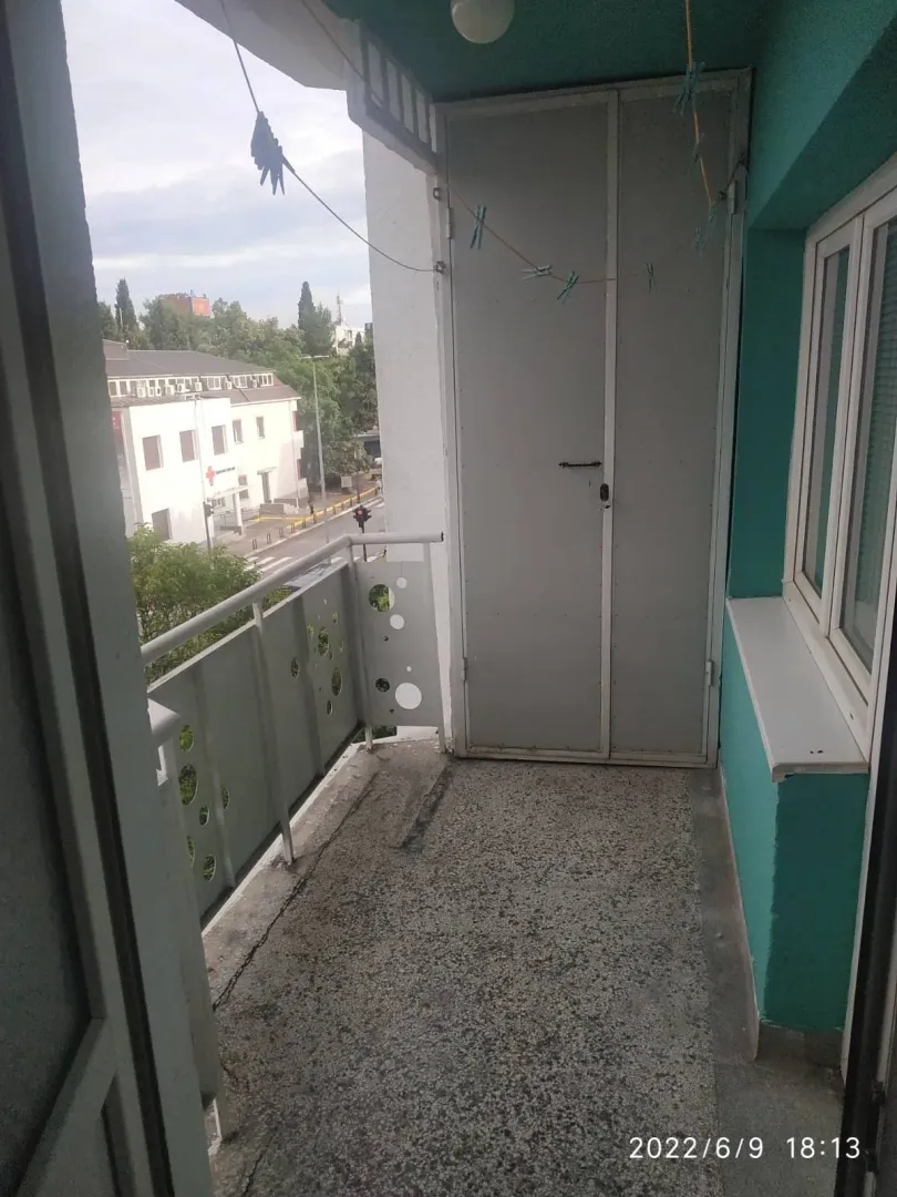 Uski balkon sa metalnom ogradom, ostavom i pogledom na ulicu.