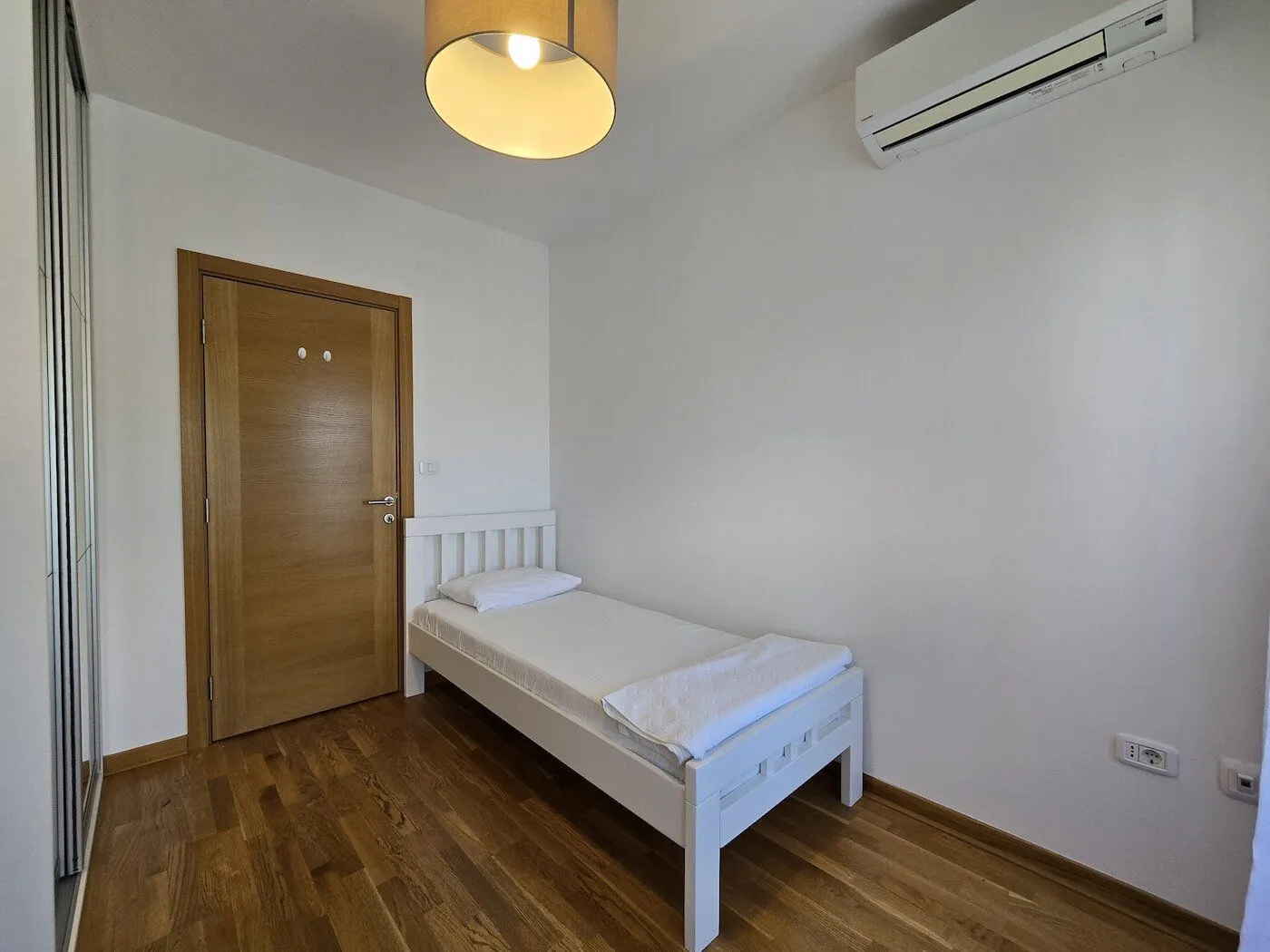 Manja spavaća soba sa bijelim krevetom, drvenim podom i klima uređajem.