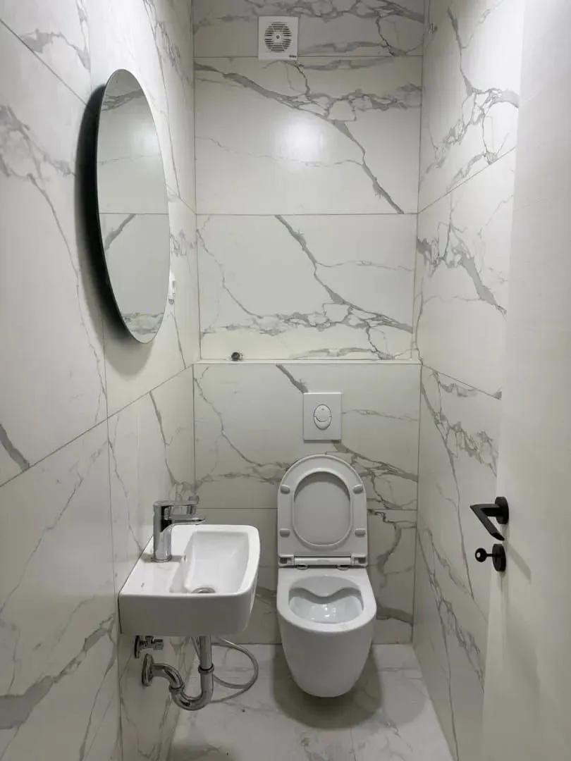 Small bathroom with white marble tiles, toilet, washbasin, and a round mirror.