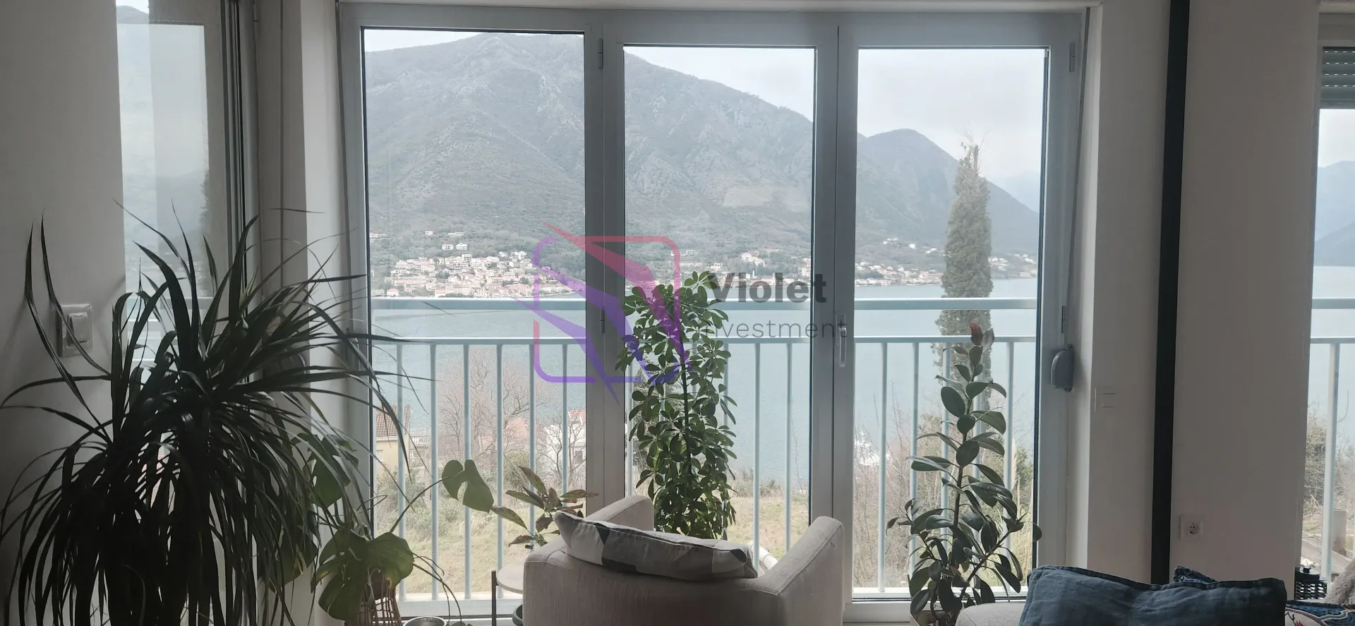 Pogled kroz velika balkonska vrata na more i planine iz dnevne sobe sa sobnim biljkama.