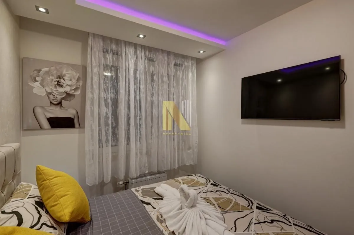 Spavaća soba sa bračnim krevetom, zidnim televizorom i dekorativnim osvetljenjem.
