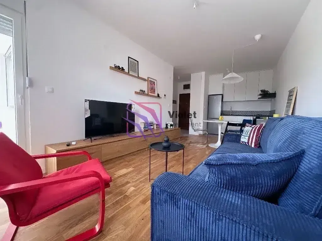 Dnevni boravak sa garniturom, televizorom i kuhinjom u pozadini.