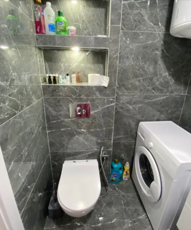 Kupatilo obloženo sivim mermernim pločicama sa WC šoljom i veš mašinom.