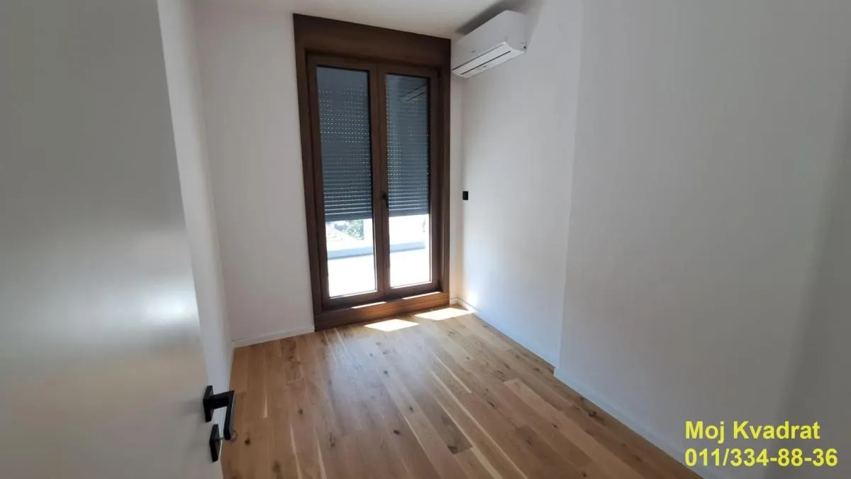 Prazna soba sa drvenim podom, balkonskim vratima i klima uređajem.