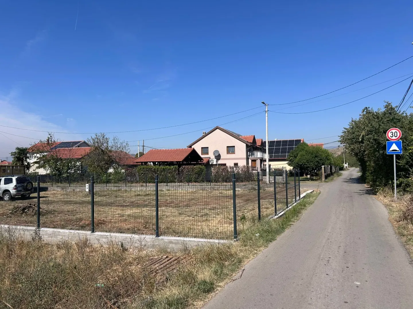 Pogled na ograđeni plac i okolne kuće pored puta.