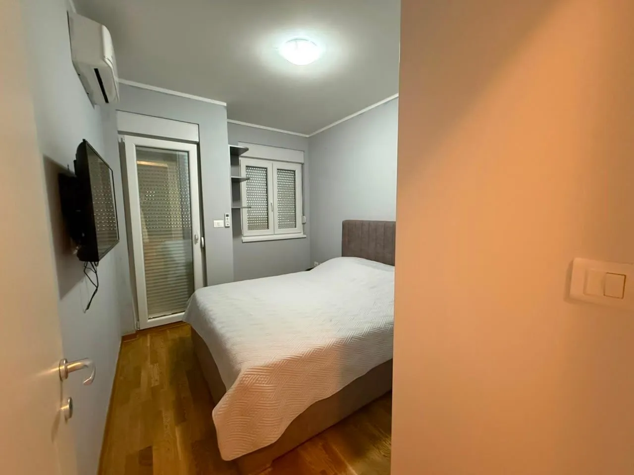 Pogled na spavaću sobu sa bračnim krevetom, televizorom na zidu i izlazom na terasu.