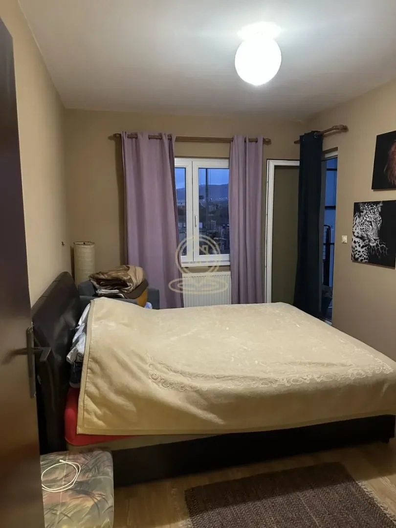 Spavaća soba sa bračnim krevetom, prozorom sa ljubičastim zavesama i slikama na zidu.