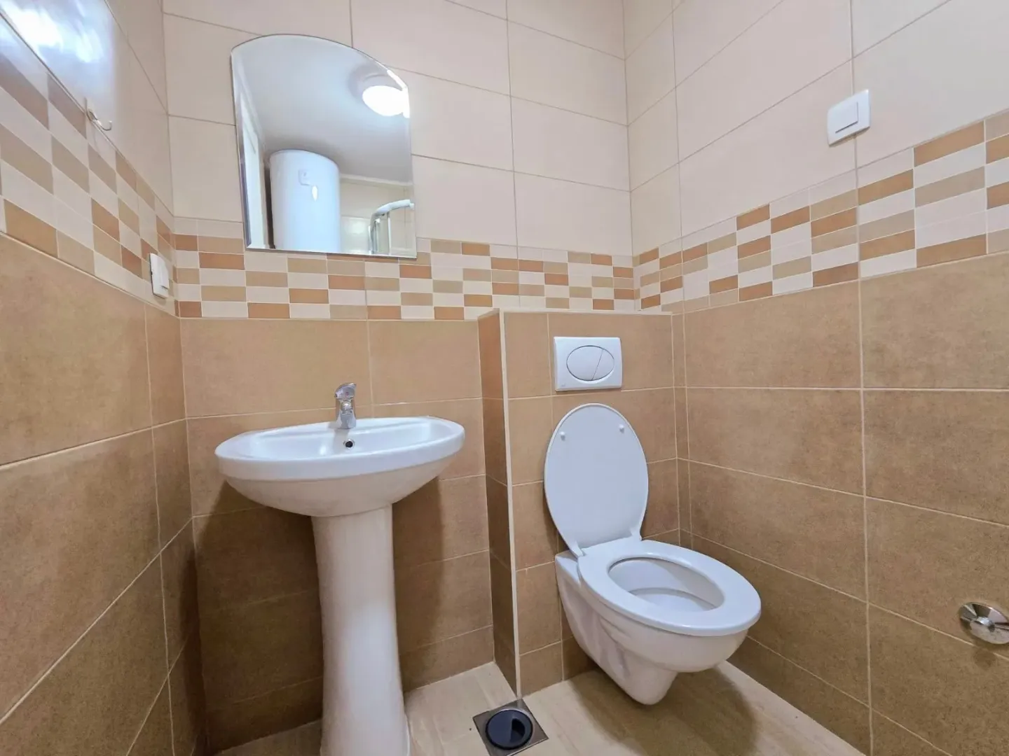 Manje kupatilo sa umivaonikom, ogledalom i wc šoljom obloženo keramičkim pločicama u bež tonovima.