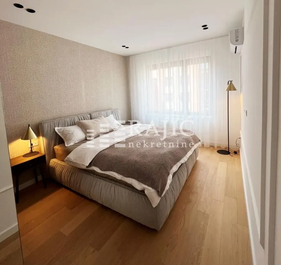 A furnished bedroom with a double bed, wooden flooring, and a window with white curtains.