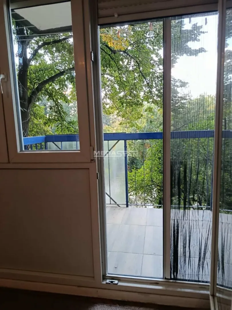 Pogled kroz balkonska vrata na terasu i zelene krošnje drveća.