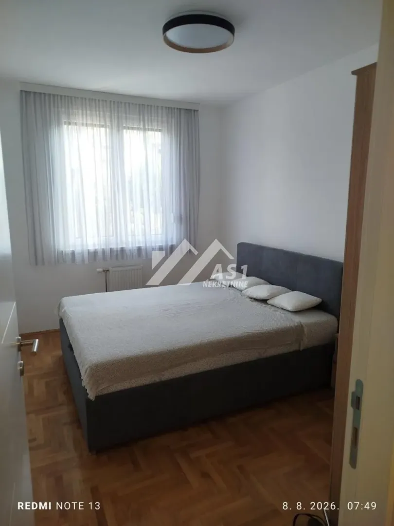 Spavaća soba sa bračnim krevetom, parketom i prozorom sa zavesama.