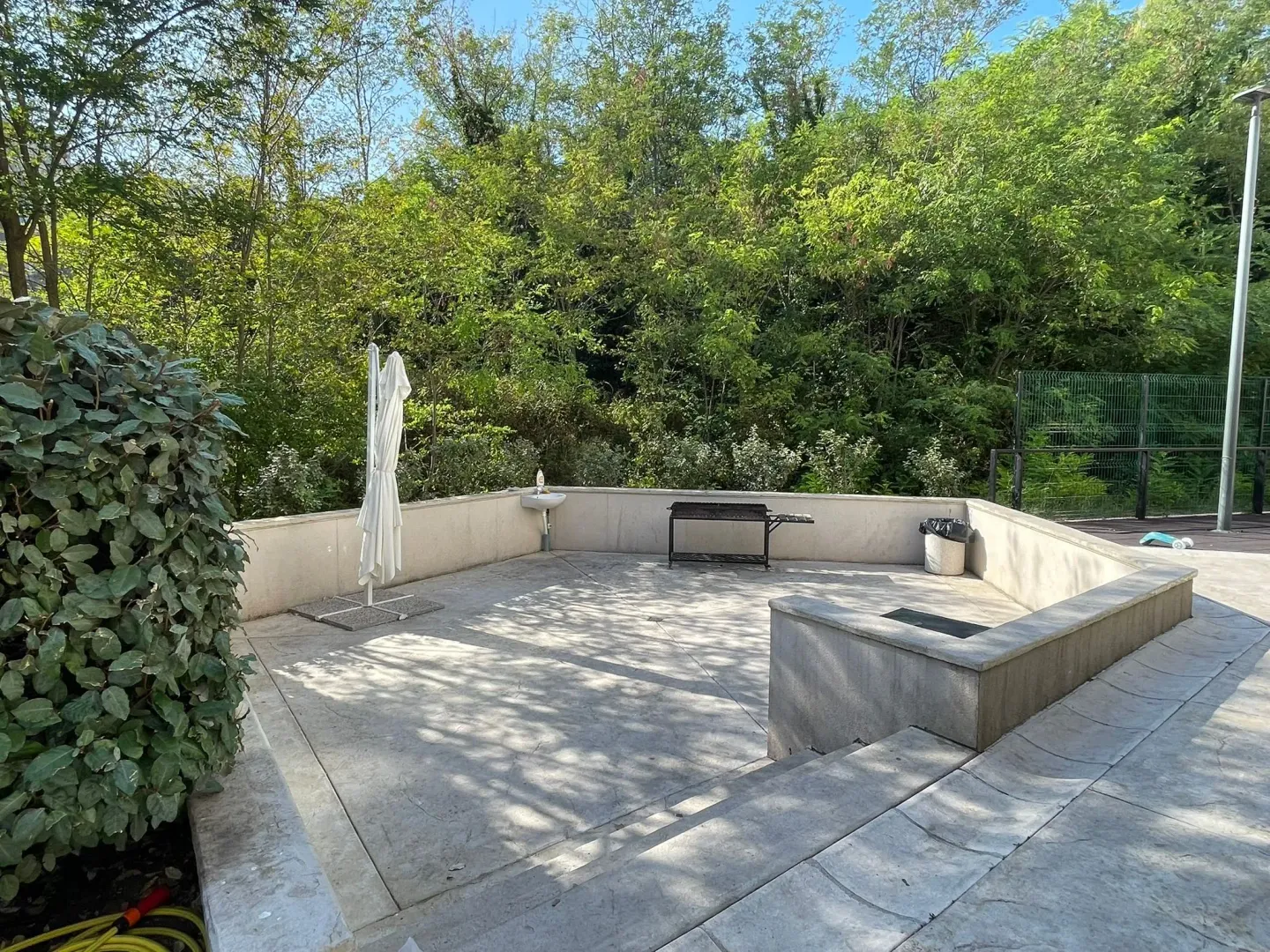 Paved yard with a barbecue, sink, and umbrella surrounded by greenery.