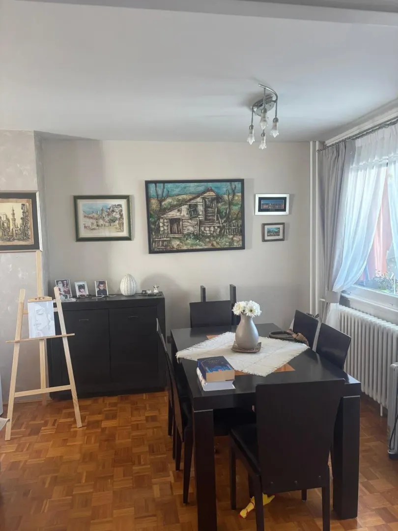 Trpezarija sa tamnim stolom i stolicama, dekorisana slikama na zidu i vazom sa cvećem.