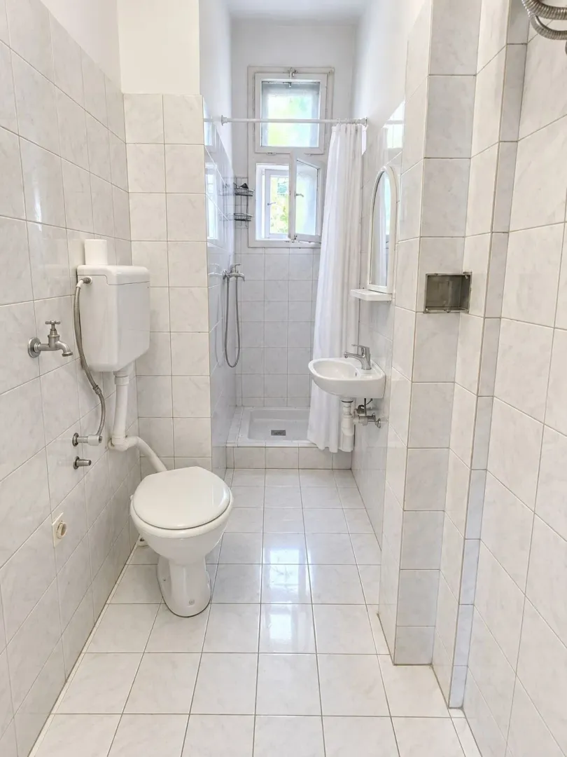 Manje kupatilo obloženo svetlim pločicama sa wc šoljom, umivaonikom i tuš kabinom.