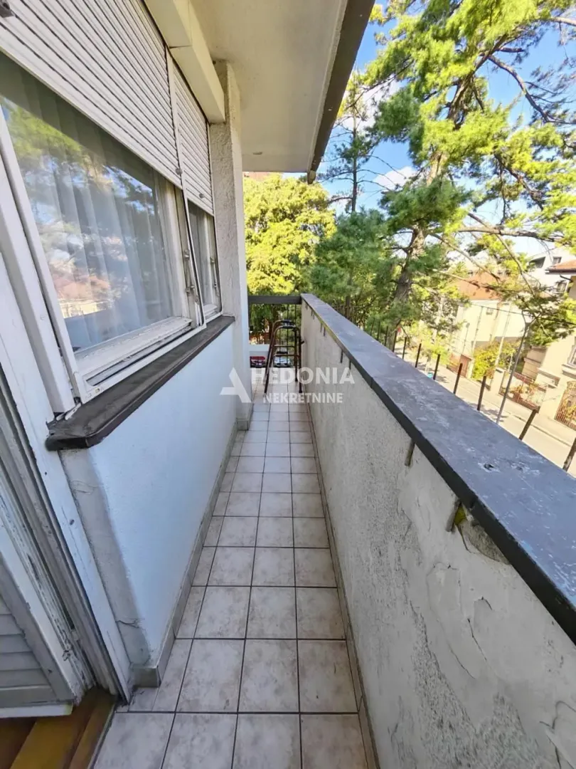 Uski popločani balkon sa pogledom na okolno zelenilo i ulicu.